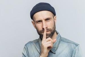 Headshot of handsome man with thick beard and mustache, has secret look, demonstrates silence sign, asks to be silent, isolated over grey background. Facial expressions and body language concept photo