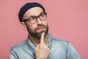 Close up shot of pleasant looking bearded male with thoughtful expression, keeps finger on chin, looks thoughtfully up, wears glasses and fashionable hat, poses against pink studio background photo