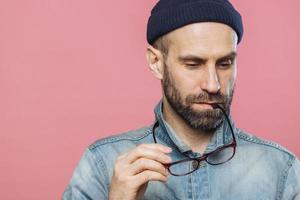 Headshot of pensive bearded man looks thoughtfully down, holds glasses, wears denim jacket and hat, isolated over pink background with copy space for your advertising content. People and thoughts photo