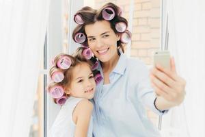 feliz madre y su hijo pequeño con rulos en la cabeza, posan para hacer selfie, usan un teléfono inteligente moderno, se paran cerca de la ventana, se preparan para las vacaciones. familia feliz, tecnología, concepto de estilo de vida foto