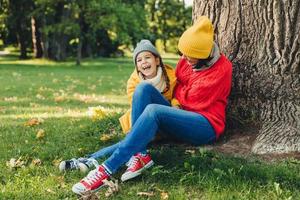 Funny little child and her mother sit together near tree, have walk in autumn park, enjoy wonderful unforgettable time together. Friendly mother and small girl, relax outdoor. Relaxation concept