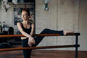 Slender smiling red haired woman stretching leg on ballet barre in classroom