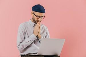 Professional male programmer keeps hans in praying gesture, looks at screen of laptop computer, believes in successful result of his work, wears formal shirt and hat, isolated over pink background photo