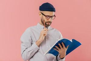 Concentrated unshaven adult man holds blue textbook and pencil, reads necessary information, has serious facial expression, wears formal clothes, isolated over pink background. Copywriter indoor photo