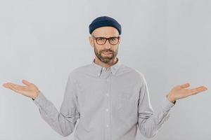 Photo of handsome man with uncertain expression, spreads hands, wears glasses and headgear, isolated over white backgrounnd. Unshaven young Caucasian male shruggs shoulders in bewilderment indoor