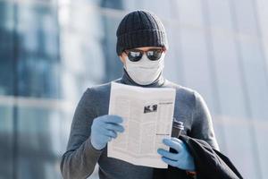 Shot of young man reads article about coronavirus symptoms, wears protective medical mask and rubber gloves, poses at street with coffee, follows rules or quarantine. Covid-19 protection concept photo