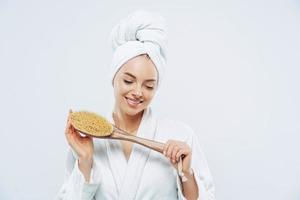 Pretty smiling woman concentrated down, holds bath brush, takes shower, cares about hygiene, wears towel on head, white soft robe, poses against white background. Women and cleanliness concept photo