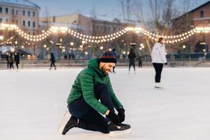 retrato de hombres alegres que atan patines para ir a patinar en la pista de hielo, preparan o se ponen zapatos especiales, disfrutan de unas maravillosas vacaciones de invierno. toma al aire libre de un encantador patinador masculino en el ring foto