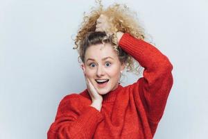 Portrait of surprised amazed happy girl with curly pony tail, stares at camera, keeps mouth widely opened, wonders her big success, models against white studio background. Excitement concept photo