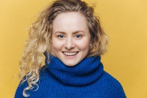 Happy delighted woman with light crisp hair, wears warm blue sweater, has broad smile and white teeth being satisfied after successful day, isolated over yellow background. Positive emotions photo