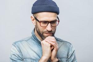 Shot of hopeful bearded male looks with thoughtful expression down, keeps hands together, hopes for better, worships in calm atmosphere, isolated over white studio background. Horizontal shot photo