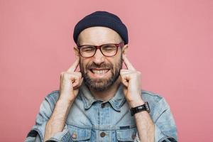 el macho barbudo molesto se tapa los oídos mientras escucha un sonido desagradable, mantiene los ojos cerrados, está insatisfecho con algo, aislado sobre fondo rosa. hombre de mediana edad sin afeitar ignora escuchar a alguien foto