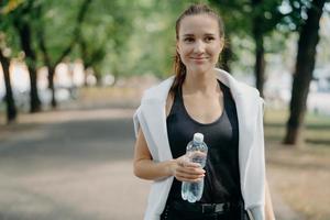 Cheerful young female runner holds water bottle poses outdoor in active wear takes break after workout walks in park gets refreshment after jogging feels thirsty keeps fit with regular sport photo
