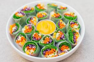 Vegan food concept. Tasty green spring rolls made of rice paper and spinach, filled with fresh chopped vegetables. Small plate with yellow curry sauce in middle photo