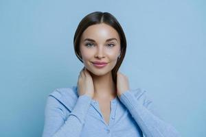 Headshot of young lovely woman keeps hands on neck, has European appearance, happy to hear pleasant words, wears casual sweater, isolated over blue background. Human face expressions concept