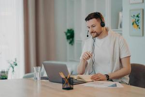 European male student learns foreign languages online has video lesson uses modern headset focused at laptop screen computer engaged in conference listens lecture attentively makes notes in diary photo