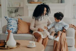 Cute little boy touching belly of his pregnant mom while relaxing together on sofa in living room photo