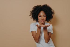 hermosa mujer africana coqueta y rizada enviando besos de aire y sonriendo mientras posa sobre fondo beige foto