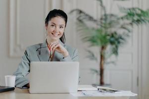 Charming stylish businesswoman works freelance at laptop computer, uses high speed internet connection, holds eyeglasses in hand, enjoys favorite remote job, reads publication, sits at office desk photo