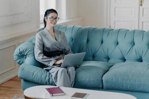 Horizontal shot of contemporary successful businesswoman poses in spacious room on comfortable sofa, reads useful article for business development, works on laptop computer, wears elegant clothes photo