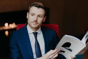 Pensive successful male employee focused into distnace, holds magazine, dressed in formal clothes, waits for business partner in cafe, thinks about signing contract and collaboration. People and work photo