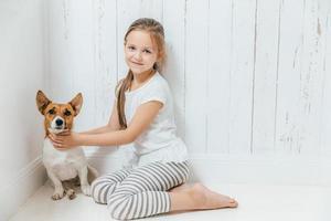 Lovely small female child plays with her dog in white room, sit on floor, have good relationship, cuddles favourite pet. Little schoolgirl likes animals, spends leisure time at home. Childhood