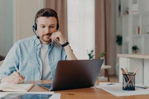 Smart man freelancer listens important info online makes necessary notes in diary works on laptop at home poses at desktop wears denim shirt poses over blurred background. Hotline support service photo