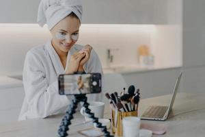 Pleased beauty blogger sits in front of cellphone camera shares experience about skin care wears hydrogel patches under eyes poses at table with cosmetic tools wears bathrobe wrapped towel on head photo