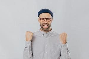 Gesture of success. Jubilant overjoyed unshaven man raises clenched fists, wears spectacles and formal shirt, celebrates his triumph, closes eyes from happiness, feels like real winner, isolated photo