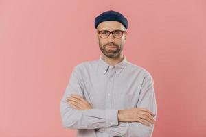 Self confident unshaven man crosses hands over chest, wears spectacles and white shirt, isolated over pink background. Bearded hipster ready for business meeting. People and fashion concept. photo