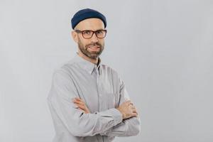 Isolated shot of pleasant looking unshaven male keeps hands crossed, wears stylish hat, optical glasses, models over white background with copy space for your advertising content, enjoys leisure photo