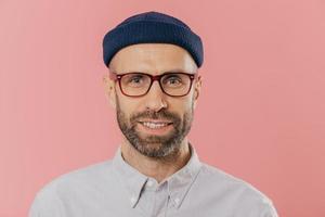 Close up portrait of smiling unshaven male, rejoices good news, wears hat and shirt, looks with eyes full of joy isolated over pink background. European man with bristle ready for date with girlfriend photo