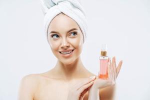 Beauty portrait of happy smiling European woman with healthy skin, makeup, holds bottle of aromatic parfume, stands naked indoor, wears towel on head. Women, cosmetology and skin care concept photo