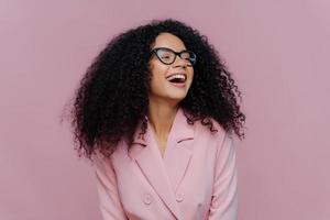 Joyous overjoyed dark skinned female manager has frizzy curly hair, laughs with happiness, wears elegant costume, transparent glasses, focused away, poses over purple background. Emotions concept photo