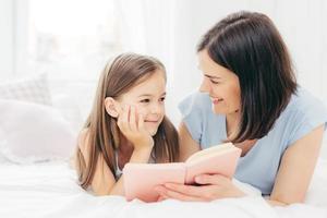 Pleased little daughter looks curiously at her mother who reads fairy tale, holds small book, lie om comfortable bed, enjoy togetherness. Cheerful small kid prepares for school. Parenthood concept photo