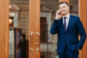 Portrait of handsome young male boss dressed in elegant expensive suit has mobile phone conversation, stands near door of cafe, rests after meeting with partners. People, business and technology photo