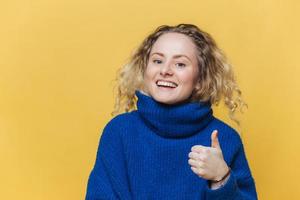 una modelo femenina positiva y de buen aspecto que está satisfecha con algo, levanta el pulgar, tiene una expresión alegre, usa un suéter azul informal, posa contra el fondo amarillo del estudio. concepto de lenguaje corporal foto