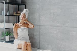 Relaxed young Caucasian female model wears towel wrapped on head, feels refreshed after taking shower, has healthy clean soft skin, poses in cozy bathroom. Women, beauty and hygiene concept. photo