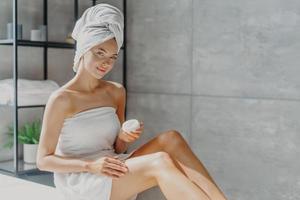 Horizontal shot of pretty woman applies cosmetic cream on skin, poses wrapped in towel, undergoes beauty procedures after taking shower, poses at home in bathroom. Daily hygiene and skin care concept photo