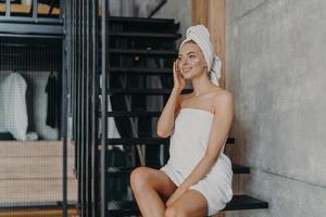 Pensive beautiful young woman applies body cream, enjoys softness of skin, sits on stairs at home, wrapped in white bath towel, looks thoughtfully into distance. Women and natural beauty concept photo