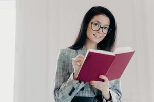 Beautiful business teacher plans her lessons, educates high school student, makes notes in notepad, wears spectacles and formal clothes, poses against white background. Woman writes down plans photo