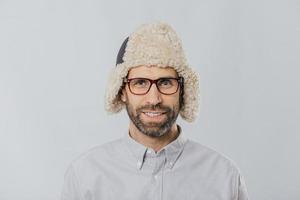 Headshot of cheerful handsome Caucasian guy wears warm winter cap with earflaps, spectacles and white shirt, looks directly at camera, ready for outdoor stroll, isolated over white background photo