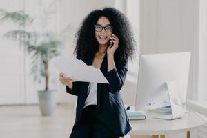 Intelligent woman with Afro hairstyle, holds paper documents, checks information, calls business partner via cell phone, analyzes future strategy, speaks with banking manager, dressed in black suit photo