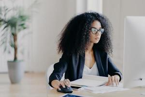 Serious curly businesswoman focused at display of computer, works on making project, surrounded with textbook and papers, wears glasses for vision correction, black suit, poses in office room photo