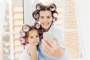 foto interior de una madre de aspecto agradable con expresión positiva y su hija hacen peinados rizados, posan para selfie en un teléfono celular moderno, se preparan para el carnaval o la fiesta. concepto de mujer y belleza