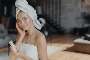 Skin care procedure concept. Adorable healthy woman applies face lotion, takes care of her skin, wrapped in bath towel, enjoys daily pampering facial treatments, has bare shoulders, healthy complexion photo