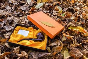 una pipa humeante en una caja de regalo yace en el suelo entre hojas amarillas de otoño foto