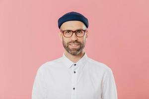 Headshot of satisfied bearded young man looks confidently at camera through spectacles, wears white formal shirt, blue hat, models against pink background, prepares for meeting with investors photo
