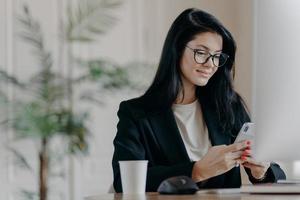 Satisfied business lady uses smartphone, poses at work place in front of computer screen, cup of tasty beverage, sends messages during break, poses at contemporary office. Technology and work concept photo