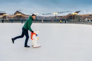 Horizontal shot of man wears gree anorak, warm hat and skates, learns to go skating on ice, uses skate aid, enjoys free time and winter holidays, smiles happily at camera. Hobby and people concept photo